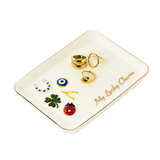 A white ceramic jewelry organizer tray with gold trim, featuring the text 'My Lucky Charm' and displaying several small charms, including a green four-leaf clover, a red ladybug, a blue and white evil eye, and gold rings, on a solid white background.