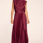 A woman modeling a sleeveless, high-neck, pleated maxi dress in a deep burgundy or plum color, standing against a plain light beige studio background.