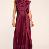 A woman modeling a sleeveless, high-neck, pleated maxi dress in a deep burgundy or plum color, standing against a plain light beige studio background.