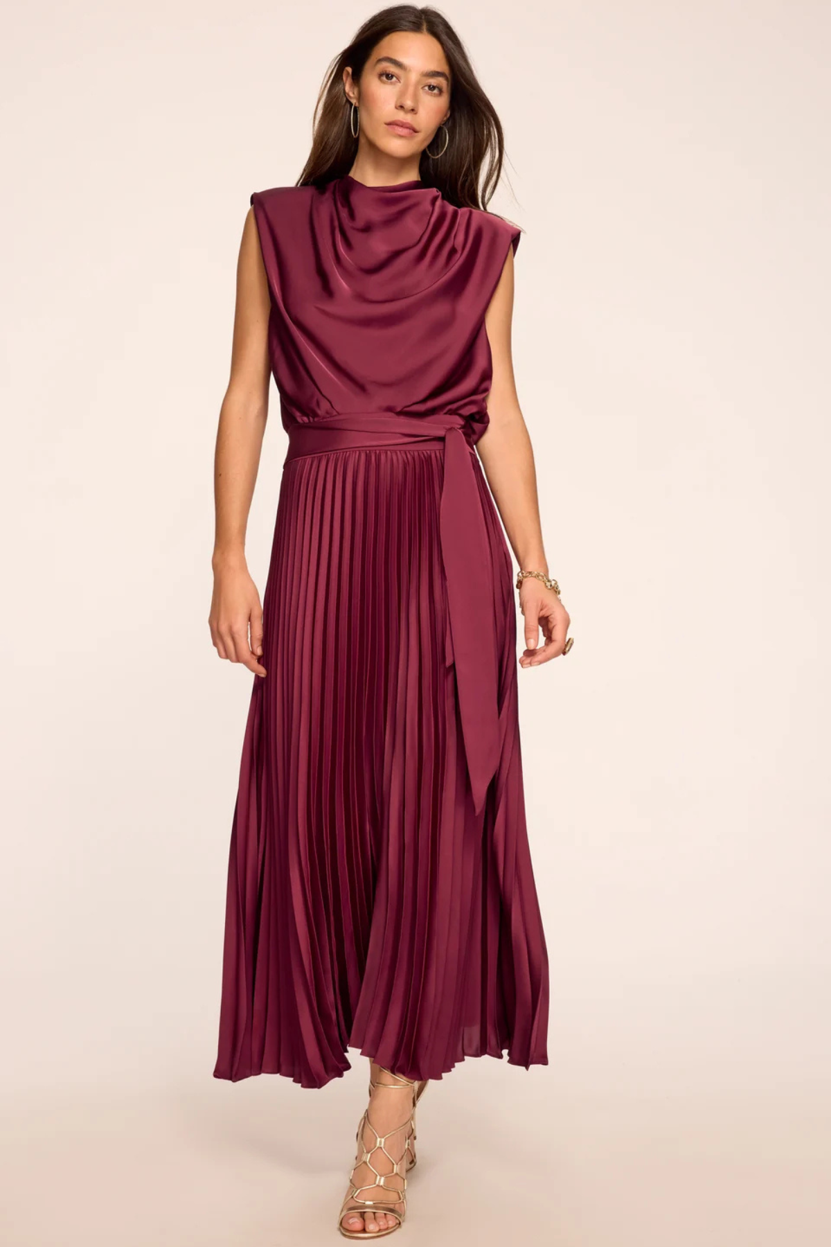A woman modeling a sleeveless, high-neck, pleated maxi dress in a deep burgundy or plum color, standing against a plain light beige studio background.