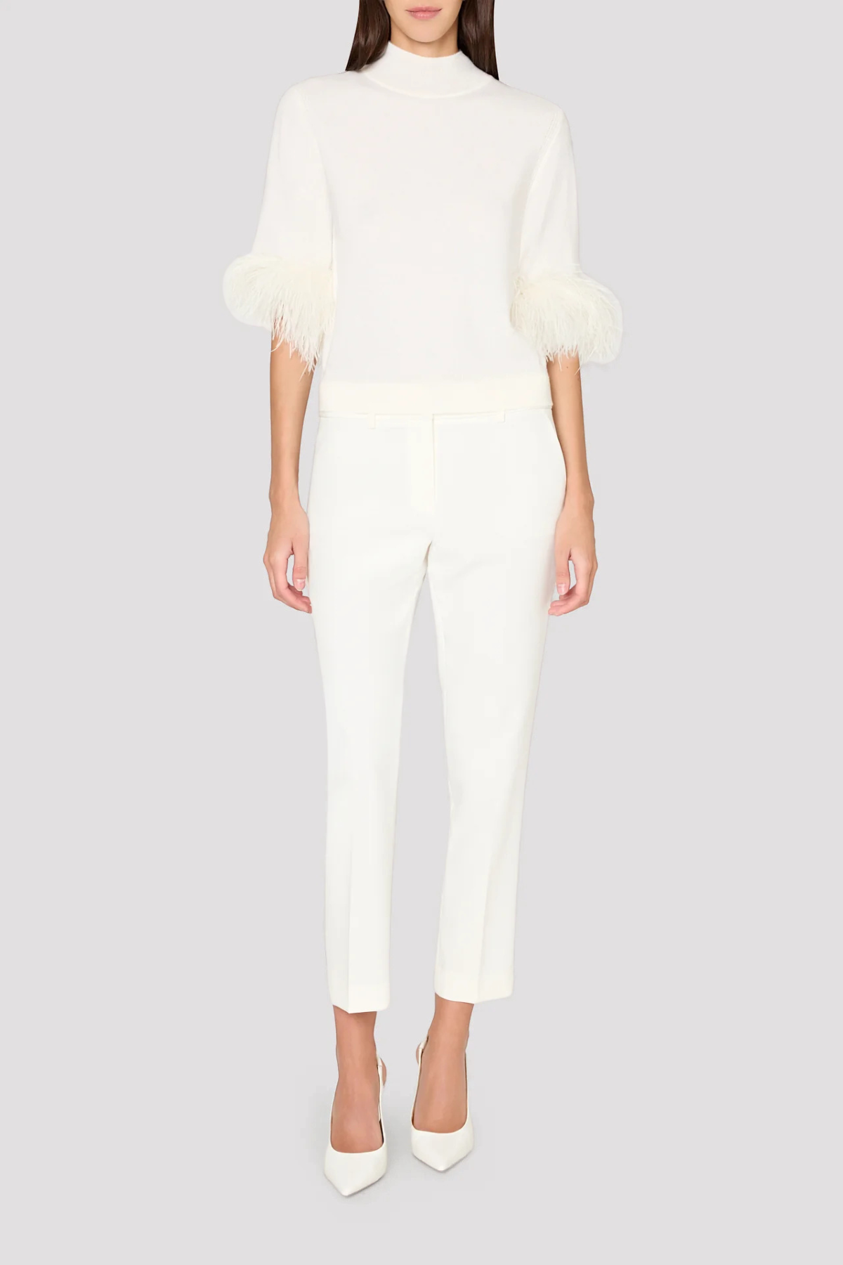 A model wearing a white mock-neck knit top with feathered elbow-length sleeves and matching white cropped trousers, paired with white pointed-toe heels, against a solid light gray background.