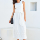 Woman wearing a white sleeveless dress with a black handbag against a white wall.