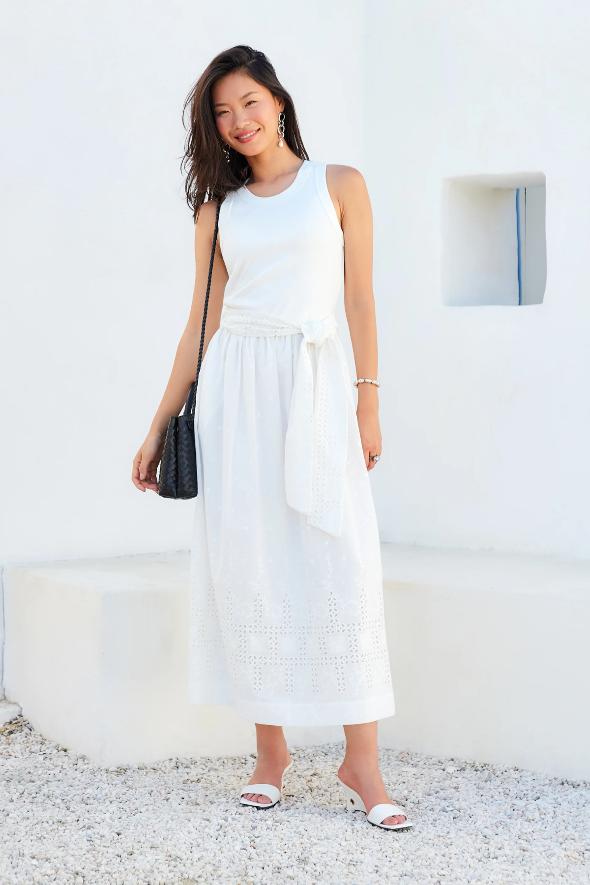 Woman wearing a white sleeveless dress with a black handbag against a white wall.