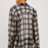 A woman shown from the back wearing a long-sleeve plaid button-up shirt in gray, black, and off-white, paired with black pants against a light gray studio background.