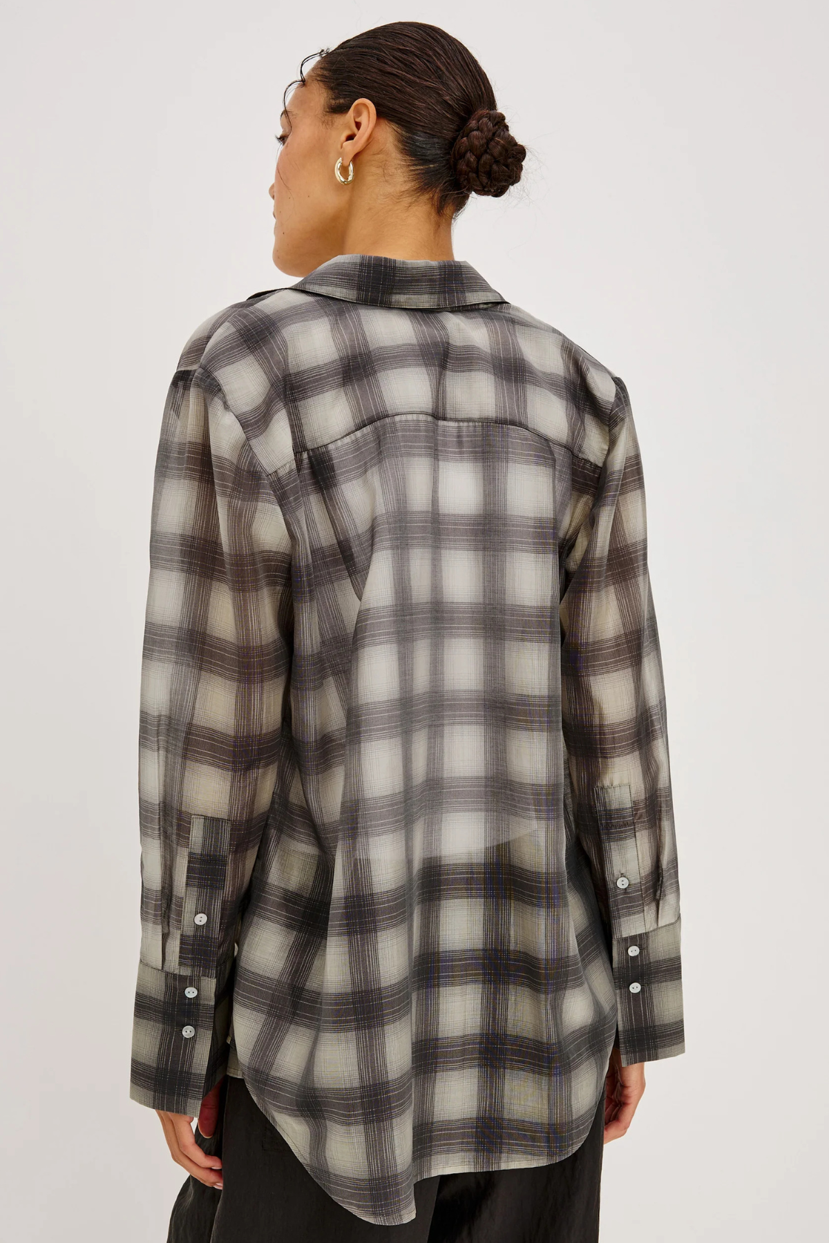 A woman shown from the back wearing a long-sleeve plaid button-up shirt in gray, black, and off-white, paired with black pants against a light gray studio background.