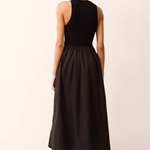 A woman shown from the back wearing a sleeveless black midi dress with a fitted bodice and flared skirt, paired with tan strappy sandals, against a plain light beige studio background.