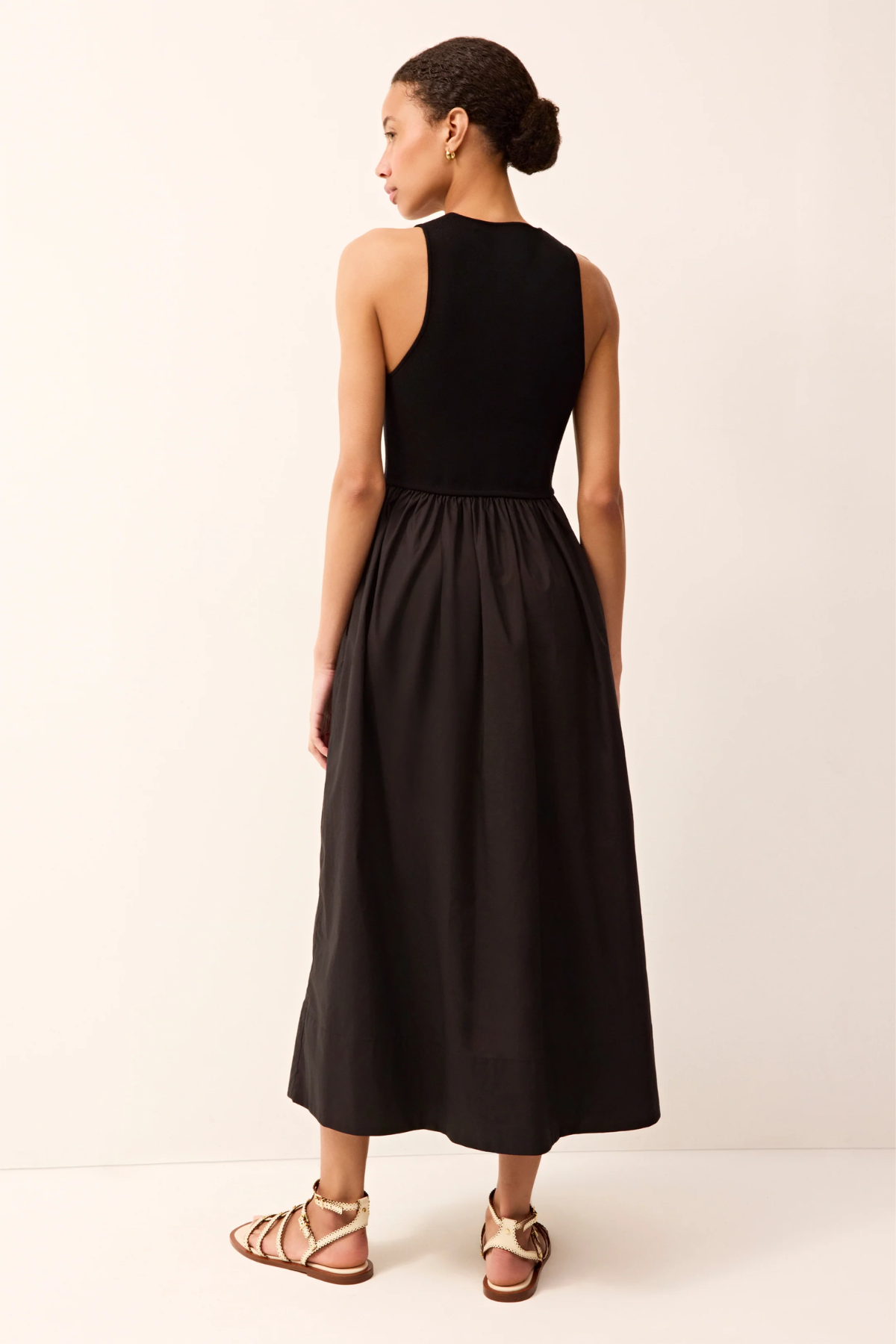 A woman shown from the back wearing a sleeveless black midi dress with a fitted bodice and flared skirt, paired with tan strappy sandals, against a plain light beige studio background.