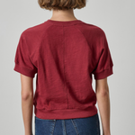A person shown from the back wearing a short-sleeve crewneck top in a muted burgundy red, paired with blue jeans, against a light gray studio background.