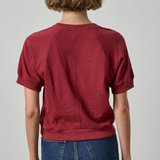A person shown from the back wearing a short-sleeve crewneck top in a muted burgundy red, paired with blue jeans, against a light gray studio background.