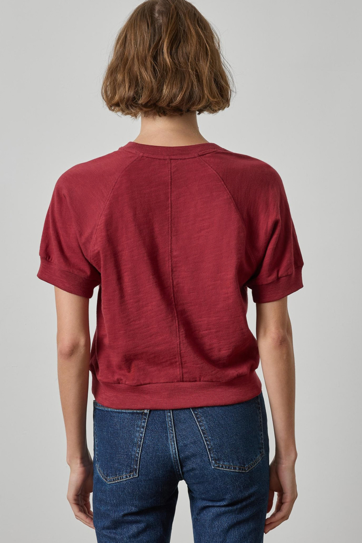 A person shown from the back wearing a short-sleeve crewneck top in a muted burgundy red, paired with blue jeans, against a light gray studio background.