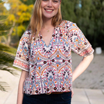 A woman wearing a short-sleeve V-neck blouse with a colorful bohemian-style geometric and floral pattern, paired with dark blue jeans, standing outdoors in front of a blurred background of trees and a patio.