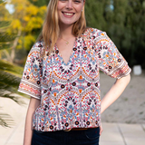 A woman wearing a short-sleeve V-neck blouse with a colorful bohemian-style geometric and floral pattern, paired with dark blue jeans, standing outdoors in front of a blurred background of trees and a patio.