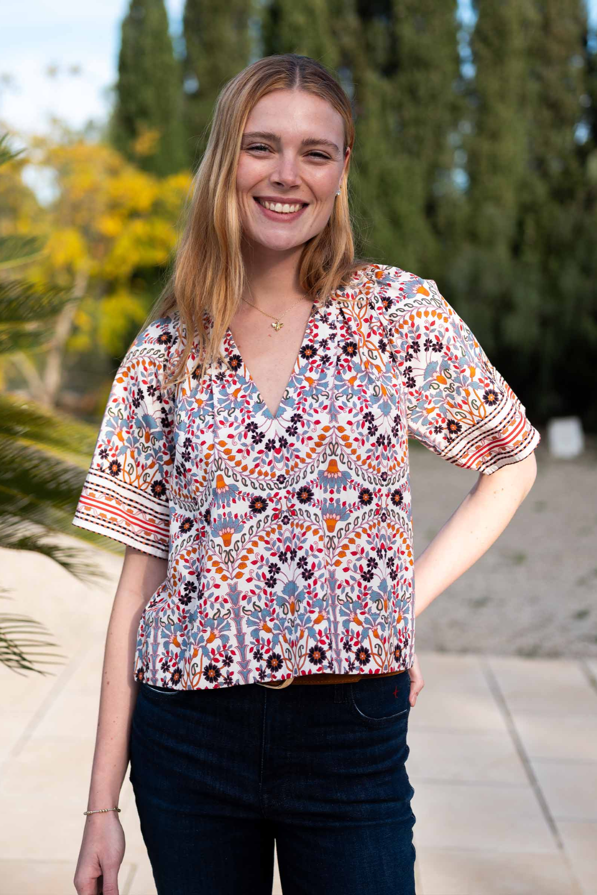 A woman wearing a short-sleeve V-neck blouse with a colorful bohemian-style geometric and floral pattern, paired with dark blue jeans, standing outdoors in front of a blurred background of trees and a patio.