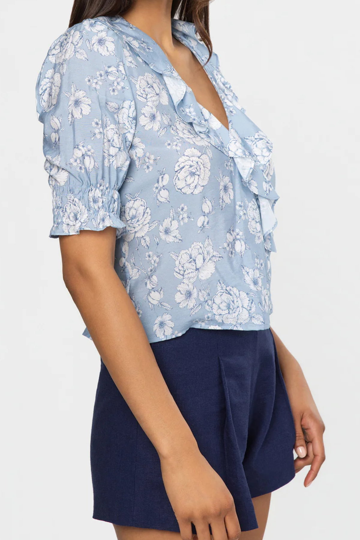 Woman wearing a light blue floral blouse with a deep neckline and navy shorts on a white background