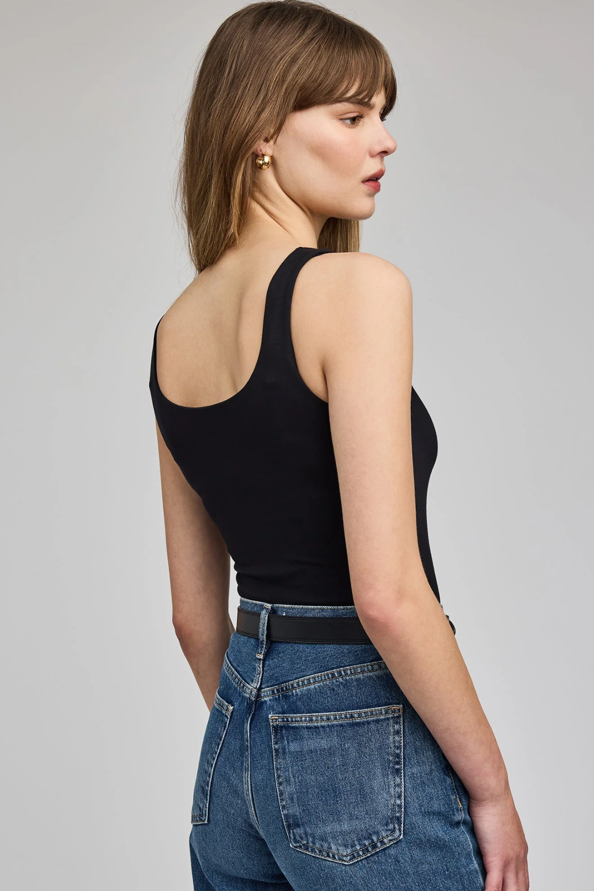 A woman shown from the back wearing a black ribbed tank top and blue jeans against a light gray studio background.