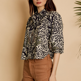 A woman modeling a black and beige botanical print blouse with a high neck and three-quarter sleeves, paired with brown trousers, set against a warm beige background with a leafy plant in the corner.