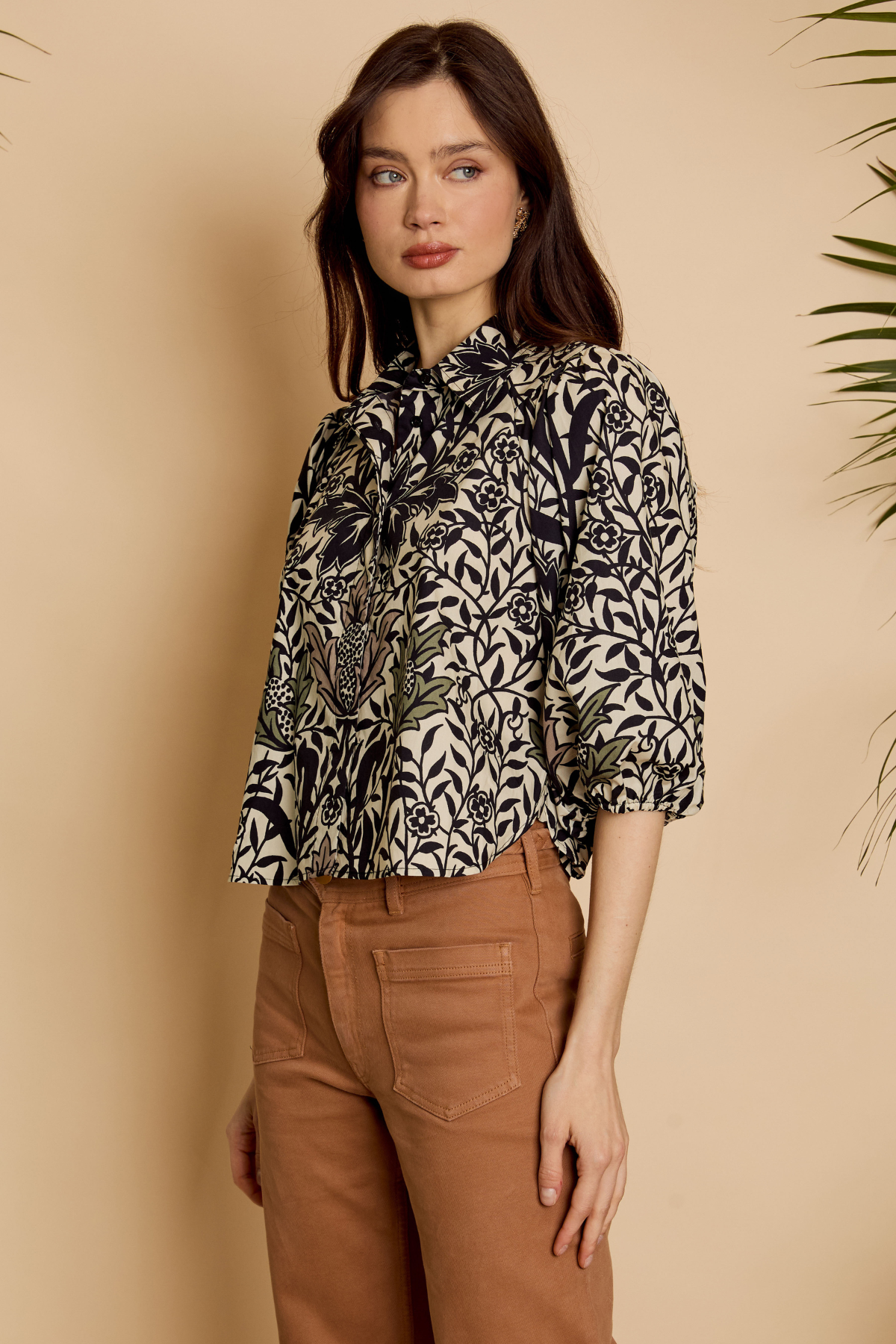 A woman modeling a black and beige botanical print blouse with a high neck and three-quarter sleeves, paired with brown trousers, set against a warm beige background with a leafy plant in the corner.