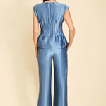 A woman viewed from the back wearing a blue satin-like sleeveless top and matching wide-leg pants against a plain beige studio background.