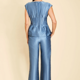 A woman viewed from the back wearing a blue satin-like sleeveless top and matching wide-leg pants against a plain beige studio background.