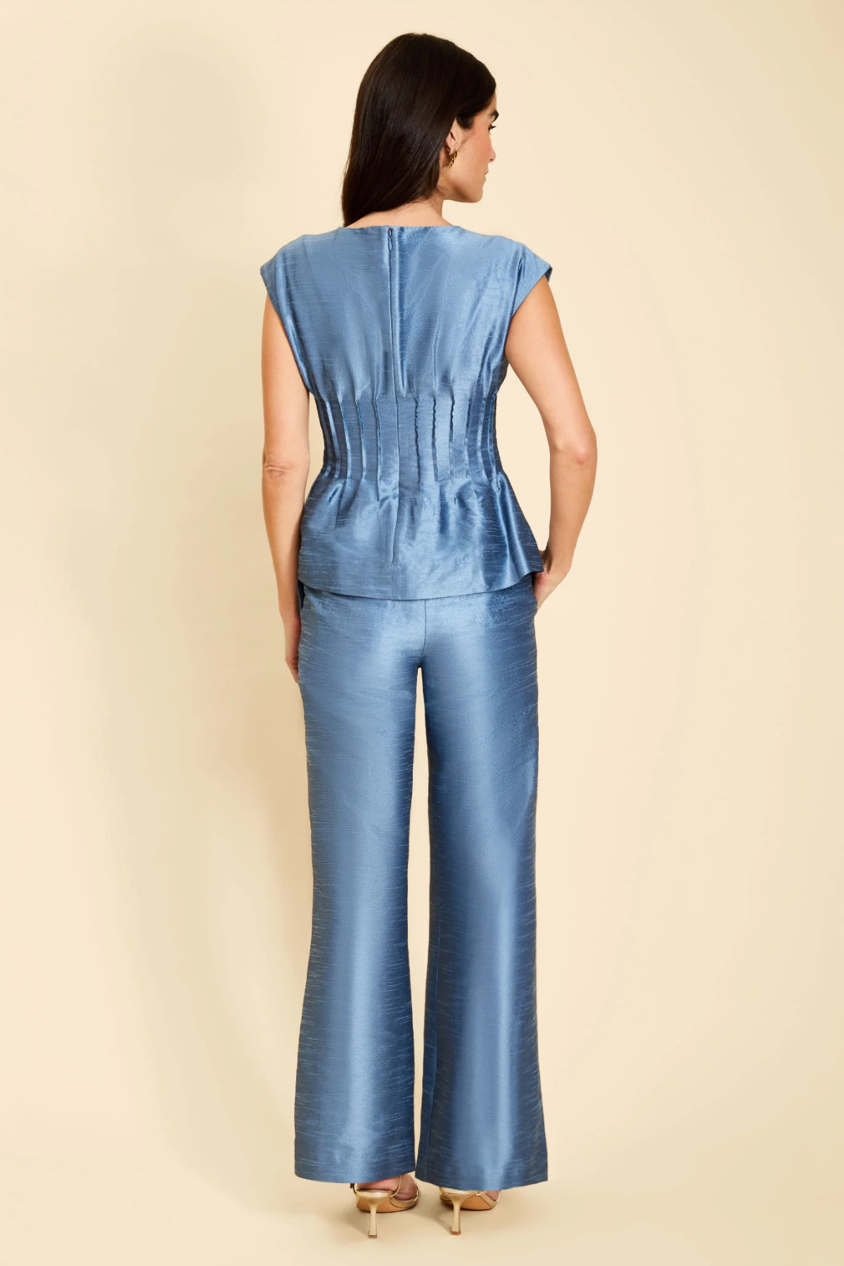 A woman viewed from the back wearing a blue satin-like sleeveless top and matching wide-leg pants against a plain beige studio background.