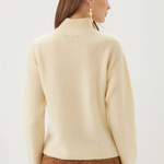 A woman shown from the back wearing a cream-colored, fuzzy mock-neck sweater and a brown pleated skirt against a plain light background.