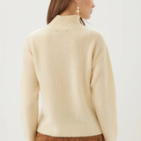 A woman shown from the back wearing a cream-colored, fuzzy mock-neck sweater and a brown pleated skirt against a plain light background.