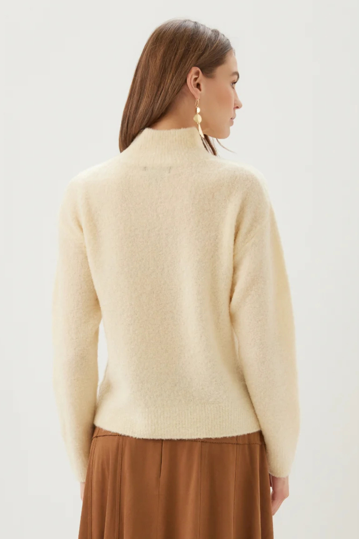 A woman shown from the back wearing a cream-colored, fuzzy mock-neck sweater and a brown pleated skirt against a plain light background.
