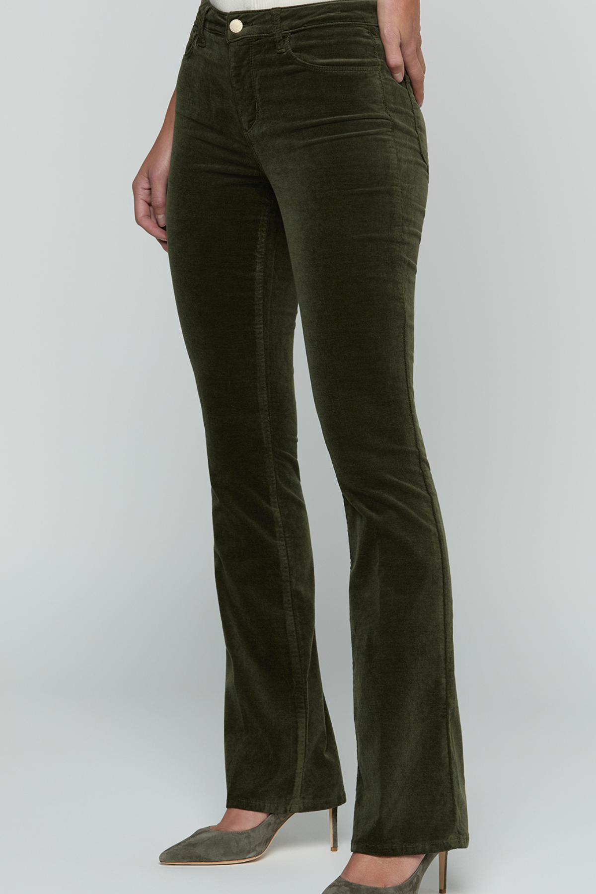 A person wearing dark olive green corduroy flare jeans with a button fly and pointed-toe heels, shown from the waist down against a light gray studio background.
