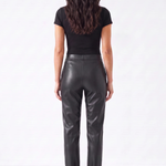 A woman viewed from the back wearing a black short-sleeve top and shiny black leather pants, standing against a plain light gray studio background.