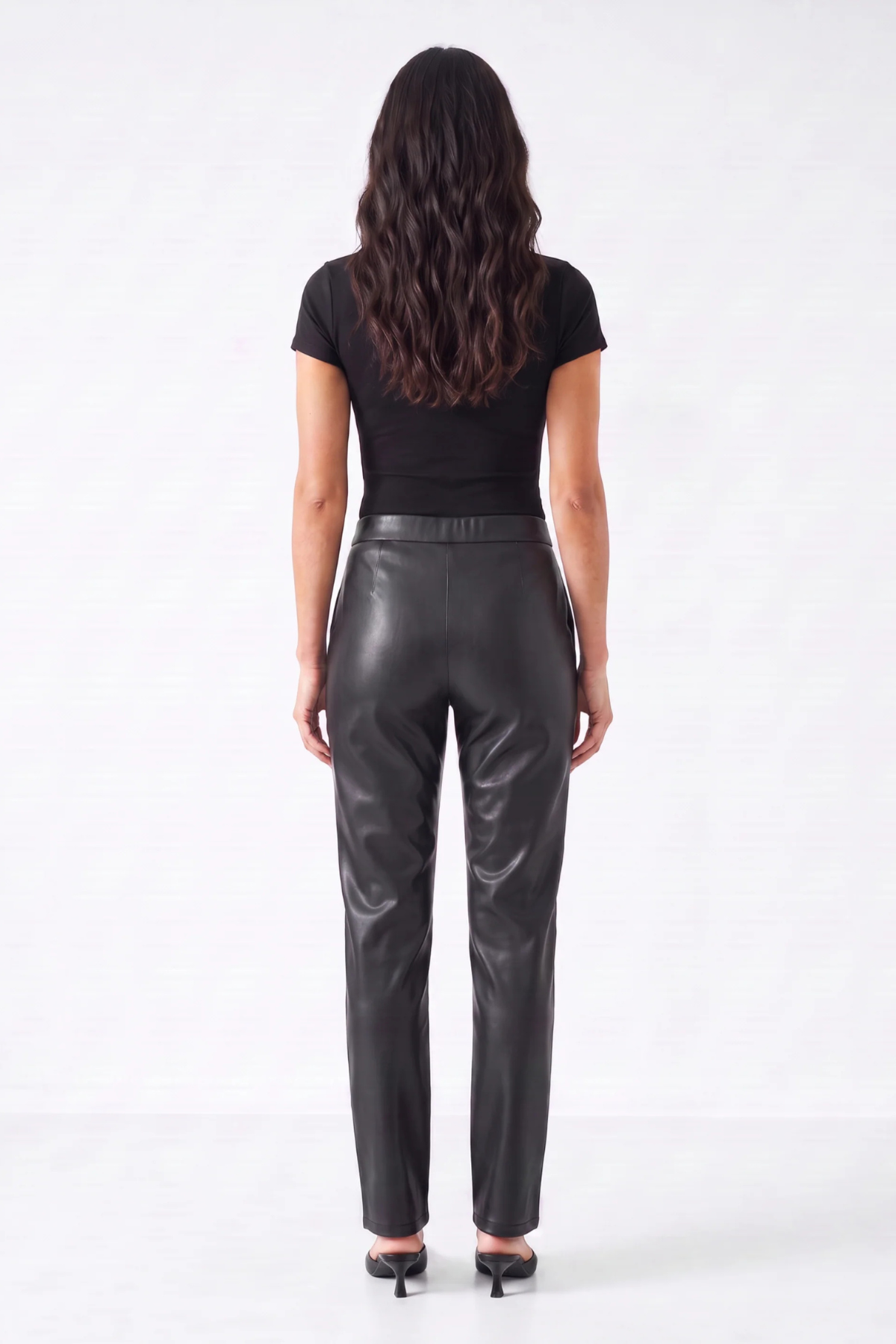 A woman viewed from the back wearing a black short-sleeve top and shiny black leather pants, standing against a plain light gray studio background.