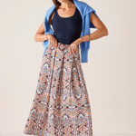 A woman modeling a long, pleated maxi skirt with a multicolor paisley pattern, styled with a navy blue tank top, a light blue cardigan, and blue Mary Jane shoes against a plain beige studio background.