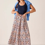 A woman modeling a long, pleated maxi skirt with a multicolor paisley pattern, styled with a navy blue tank top, a light blue cardigan, and blue Mary Jane shoes against a plain beige studio background.
