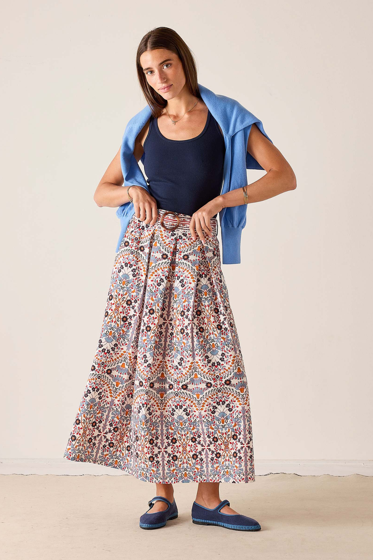 A woman modeling a long, pleated maxi skirt with a multicolor paisley pattern, styled with a navy blue tank top, a light blue cardigan, and blue Mary Jane shoes against a plain beige studio background.