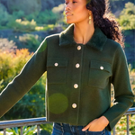 A woman wearing a dark green cropped knit jacket with a fuzzy collar and white buttons, paired with blue jeans, posing outdoors against a green hillside background.