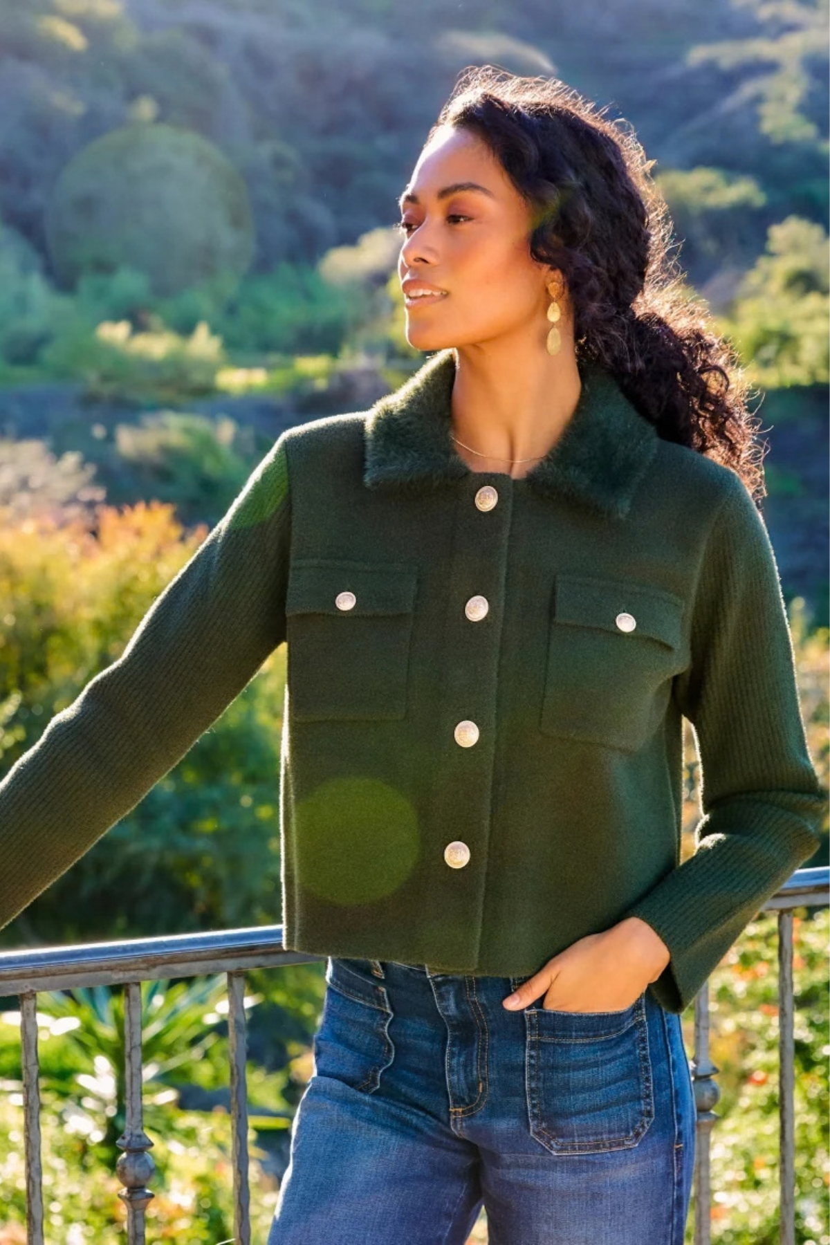 A woman wearing a dark green cropped knit jacket with a fuzzy collar and white buttons, paired with blue jeans, posing outdoors against a green hillside background.