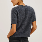 A woman shown from the back wearing a short-sleeve fuzzy knit sweater in dark charcoal gray, paired with dark jeans, against a plain light gray studio background.