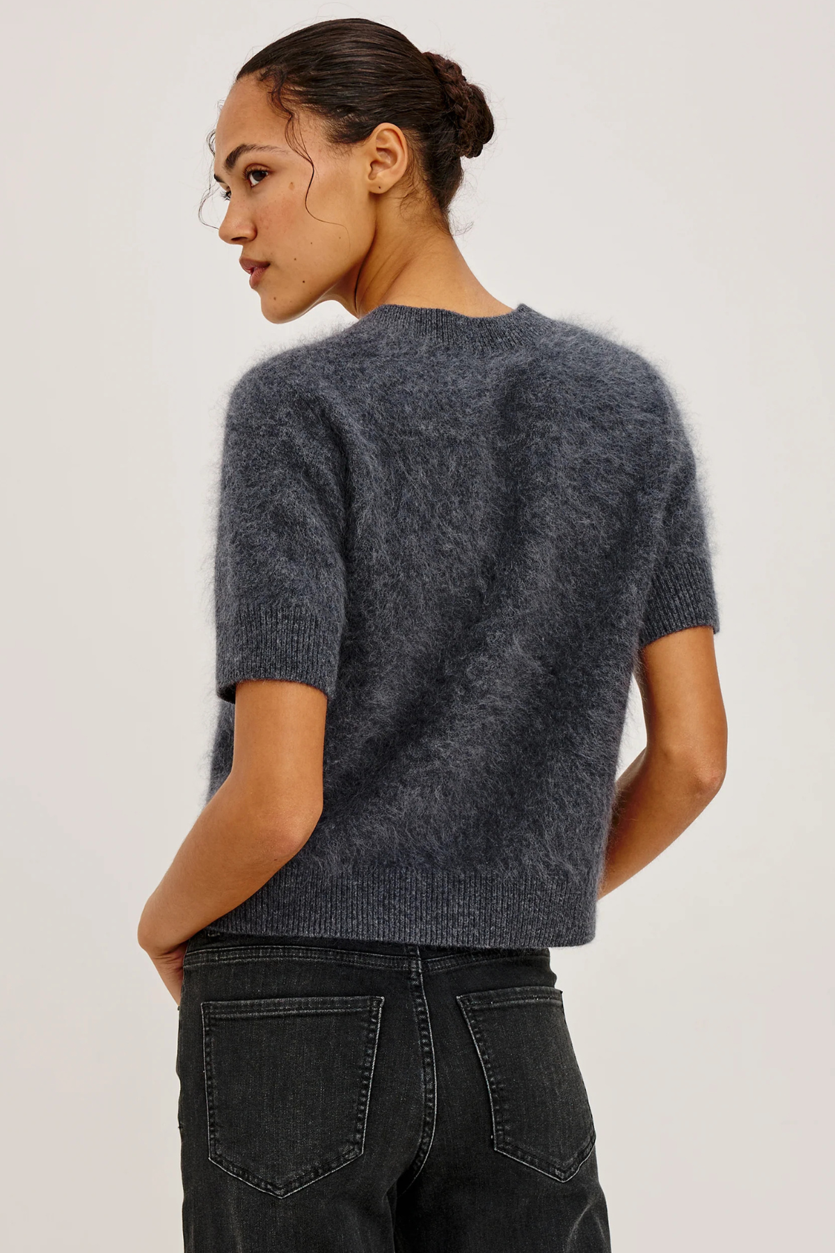 A woman shown from the back wearing a short-sleeve fuzzy knit sweater in dark charcoal gray, paired with dark jeans, against a plain light gray studio background.