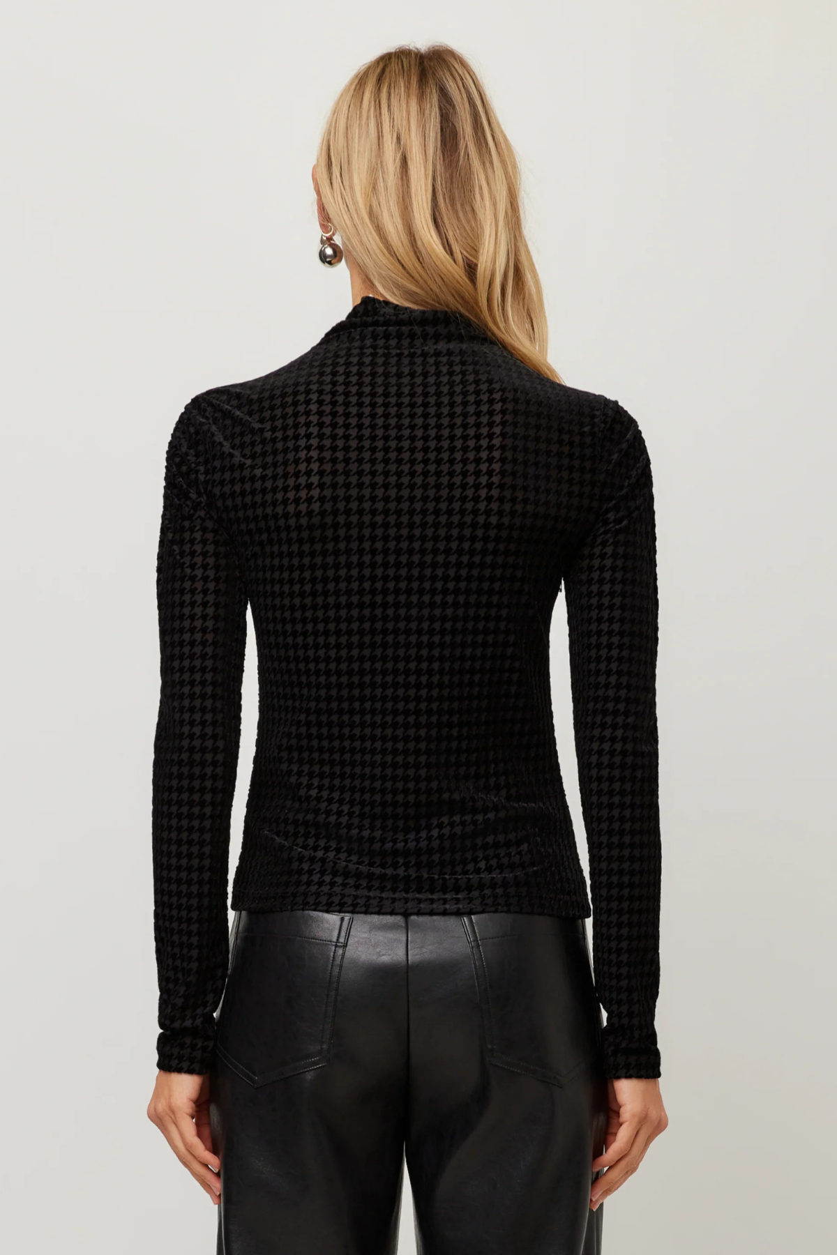 A woman viewed from the back wearing a black long-sleeve mock-neck top with a fine houndstooth pattern, paired with black leather pants, against a light gray studio background.