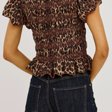 A woman wearing a leopard-print smocked top with ruffled short sleeves and a ruffled hem, paired with dark blue jeans, shown from the back against a light gray background.