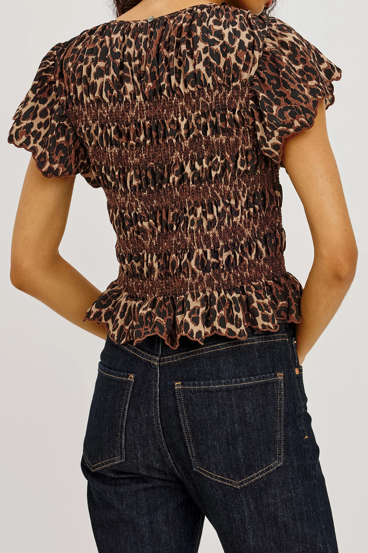 A woman wearing a leopard-print smocked top with ruffled short sleeves and a ruffled hem, paired with dark blue jeans, shown from the back against a light gray background.
