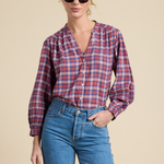 A woman modeling a long-sleeve plaid button-up shirt in red, blue, and white, paired with blue jeans and black sunglasses, against a solid beige background.