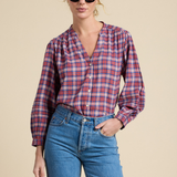 A woman modeling a long-sleeve plaid button-up shirt in red, blue, and white, paired with blue jeans and black sunglasses, against a solid beige background.
