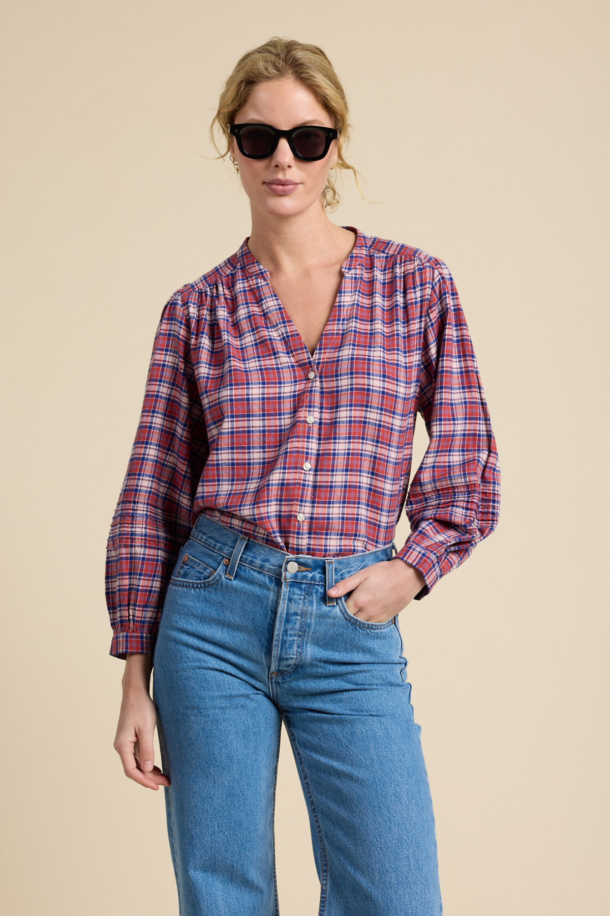 A woman modeling a long-sleeve plaid button-up shirt in red, blue, and white, paired with blue jeans and black sunglasses, against a solid beige background.