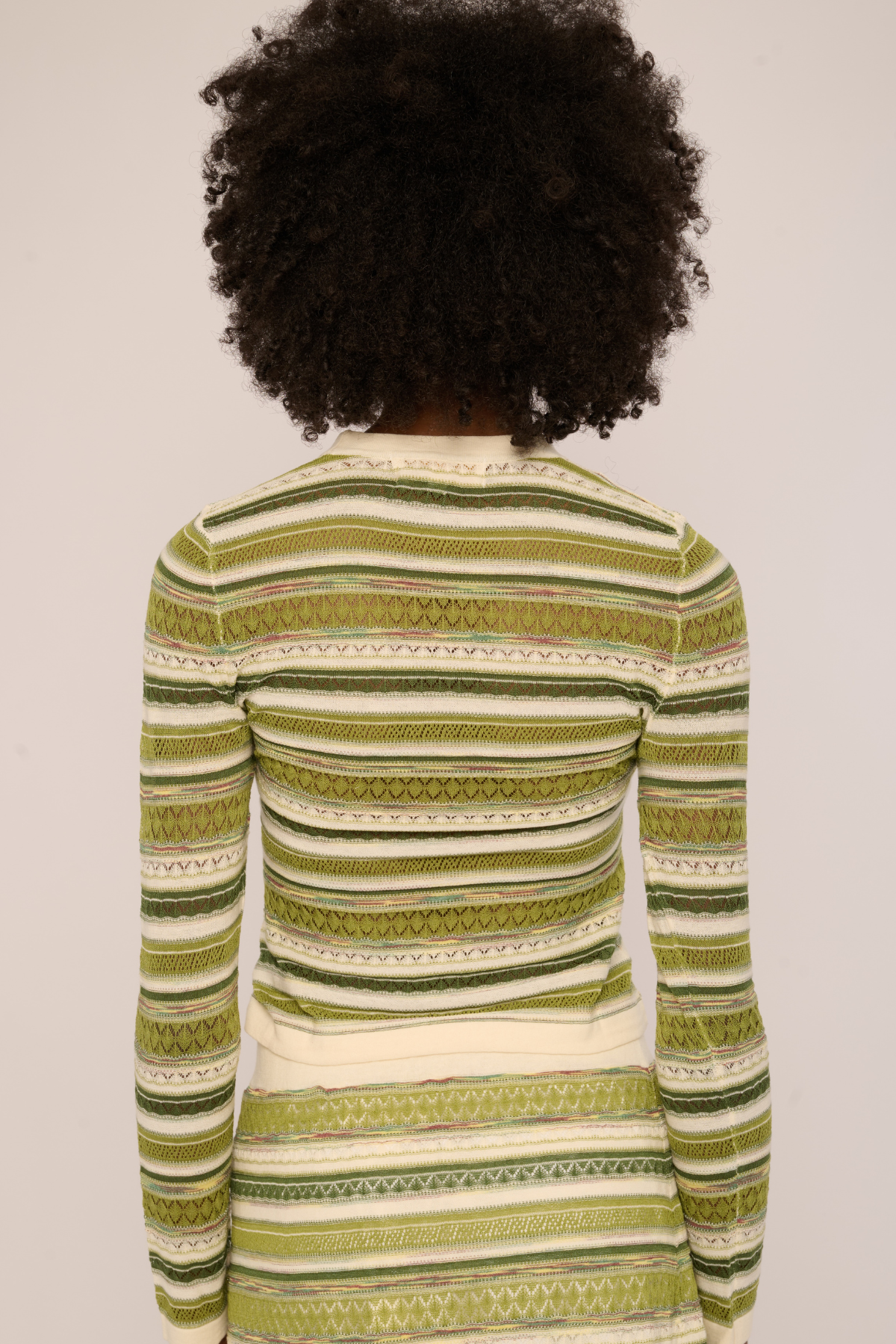 A person viewed from the back wearing a long-sleeve knit top with horizontal stripes in olive green, sage green, and cream on a light gray background.