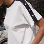 White t-shirt with black trim and silver buttons worn by a person outdoors.