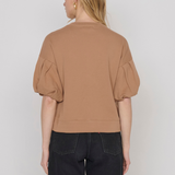 A woman shown from the back wearing a cropped tan top with voluminous puff sleeves and black jeans, photographed against a plain light gray studio background.