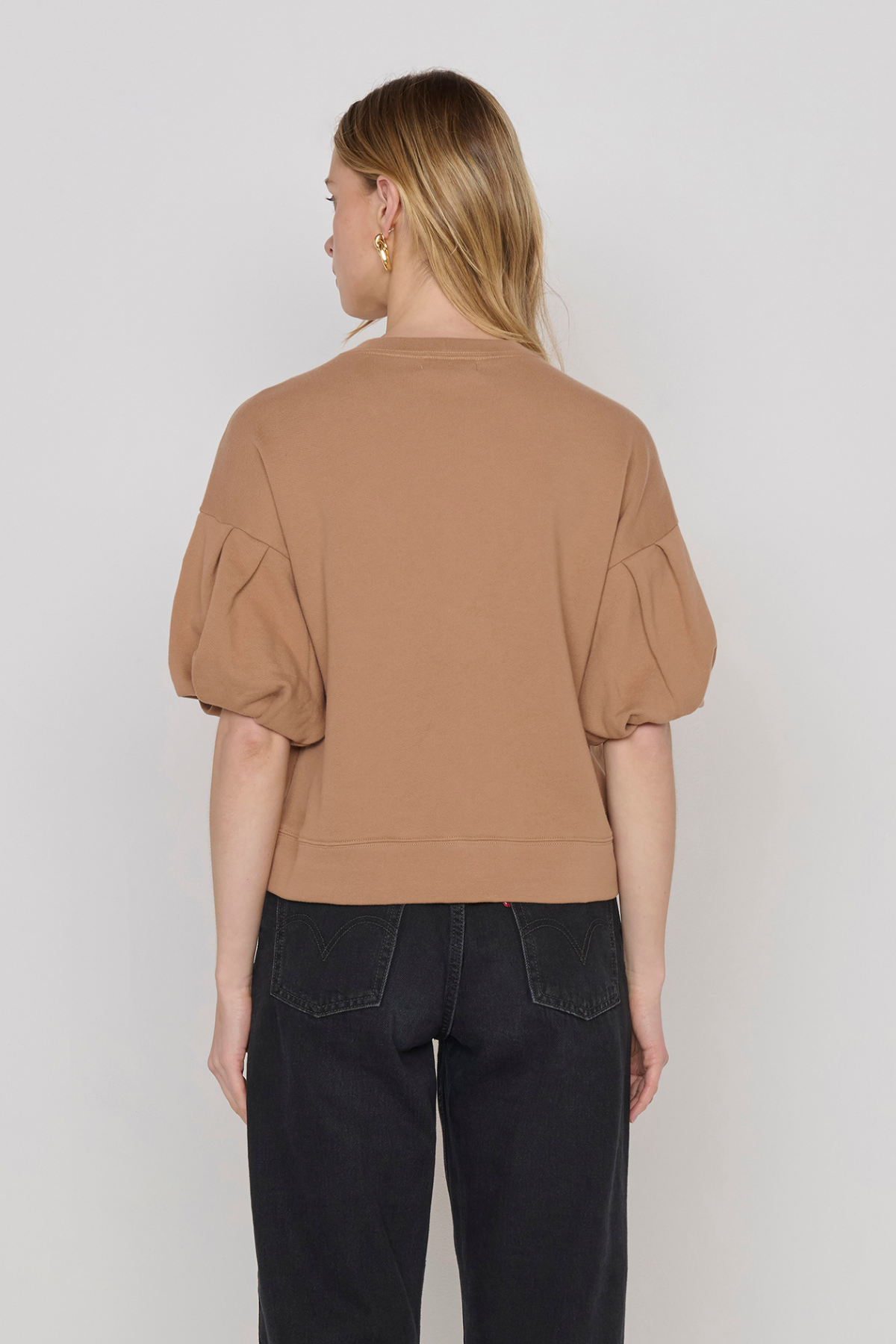 A woman shown from the back wearing a cropped tan top with voluminous puff sleeves and black jeans, photographed against a plain light gray studio background.