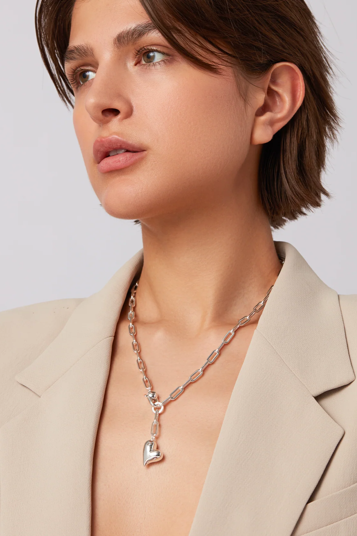 Woman wearing a silver necklace with a heart pendant against a plain background