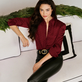 A woman posing on a white sofa wearing a deep burgundy textured long-sleeve blouse and black faux-leather pants with a gold-buckled belt.