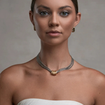 A woman with her hair pulled back wears a delicate silver chain necklace with a gold-toned textured center bead. She is wearing a white strapless top against a neutral gray studio background.
